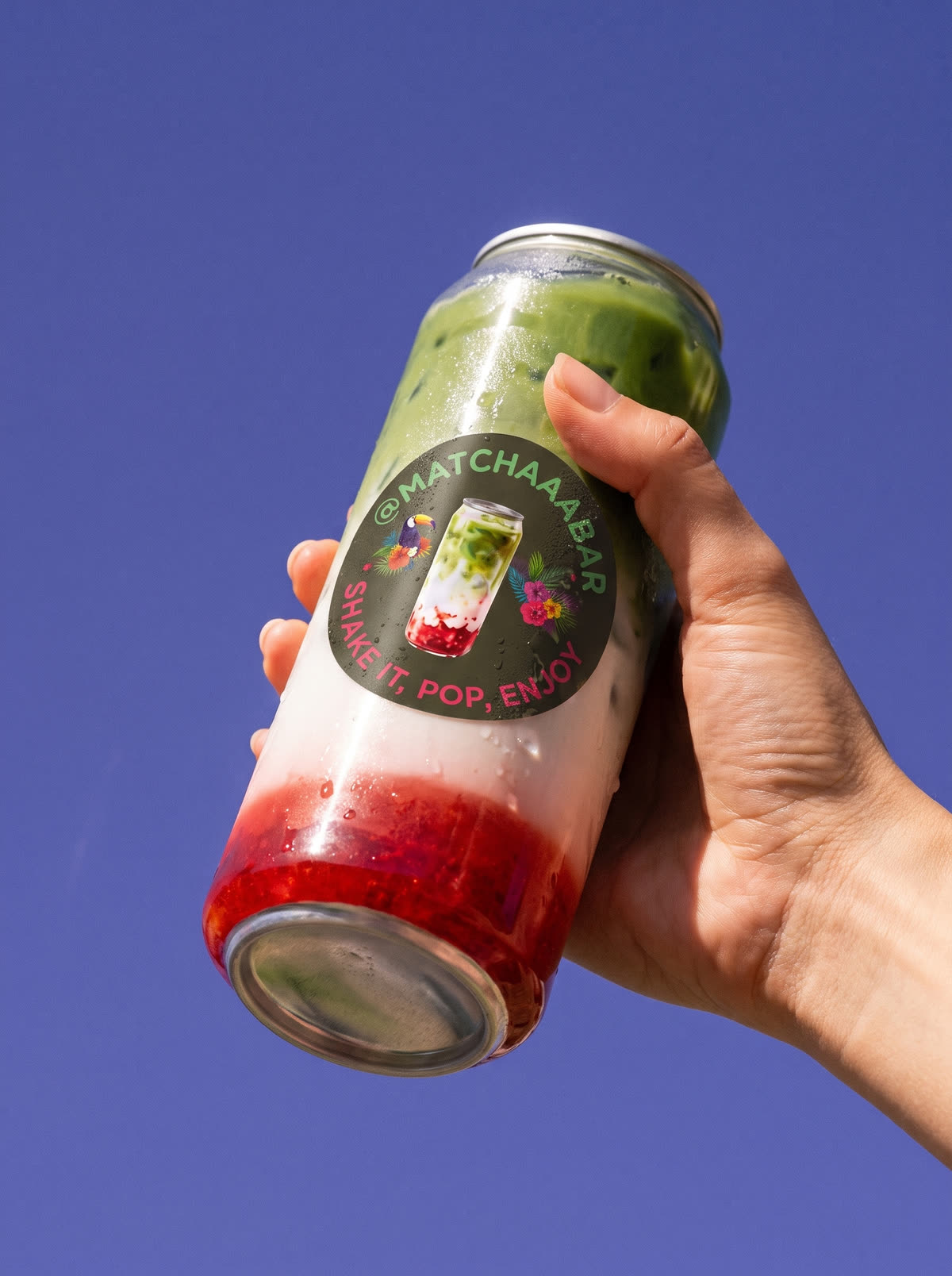 Een hand houdt een matcha van @matchaaabar omhoog tegen een blauwe lucht. In het flesje liggen de lagen op elkaar: fruit onderin, melk in het midden en matcha bovenop.